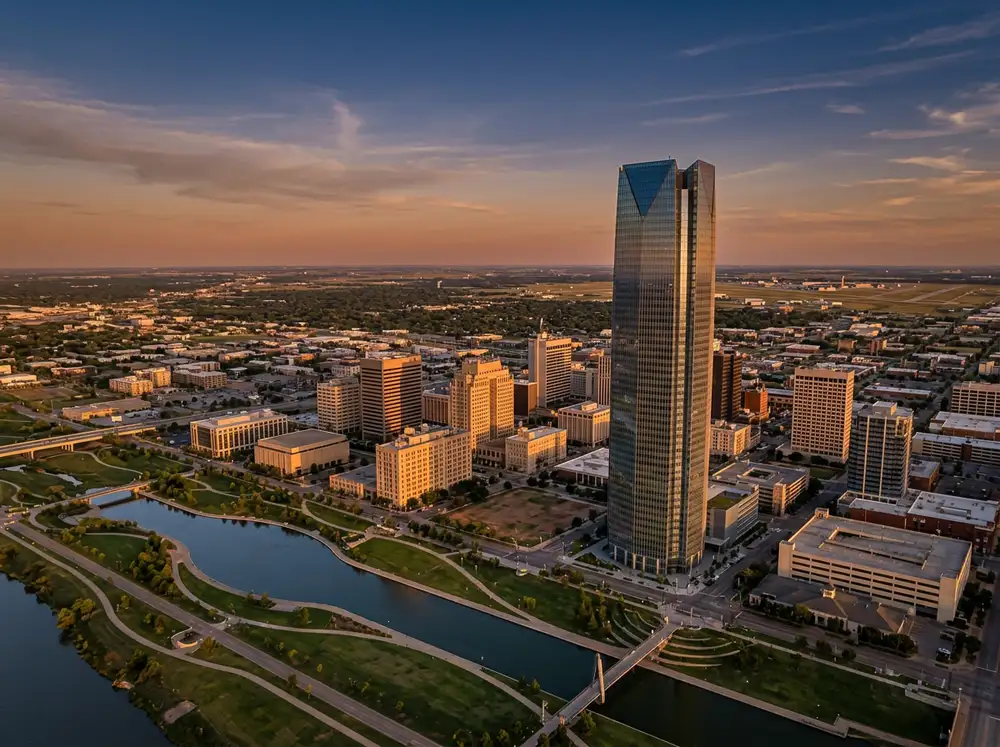 Aerial daytime view of Oklahoma City at golden hour showing the Devon Tower glass facade glowing in warm sunset light Scissortail Park below and the suburban prairie grid stretching east toward Tinker Air Force Base