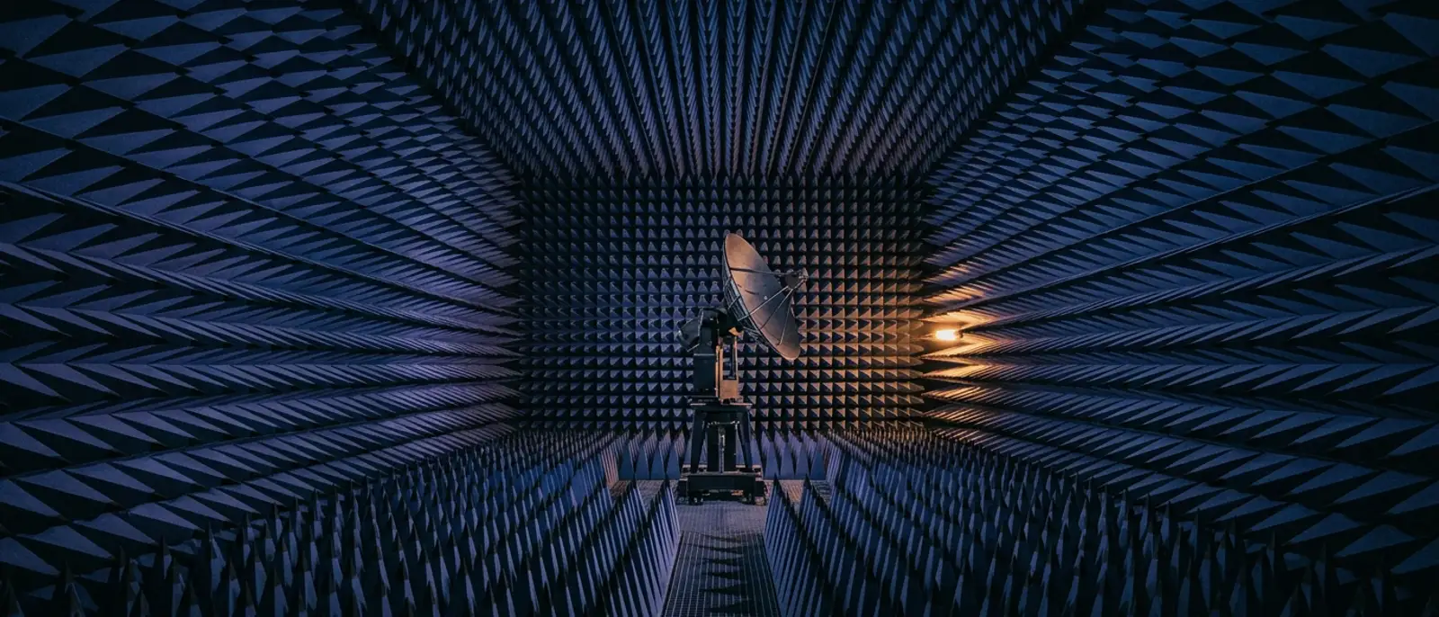 Wide view down a microwave anechoic chamber lined with blue pyramidal radio-absorbing foam with a parabolic test antenna on a positioner