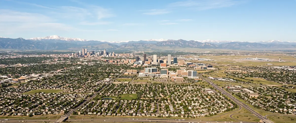 High aerial daytime view of Aurora Colorado stretching east across the high plains from the Anschutz Medical Campus toward Buckley Space Force Base and Denver International Airport, with the snow-capped Front Range and Rocky Mountains rising on the western horizon