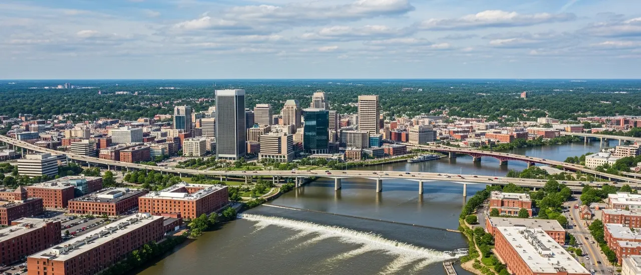 High aerial daytime view of the Richmond Virginia region along the James River, with the downtown skyline, the rapids of the fall line, historic brick warehouses on the riverbank, the I-95 bridges crossing the water, and the green suburbs of Henrico and Chesterfield under a clear sky