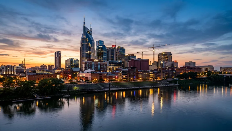The downtown Nashville skyline at dusk with the AT&T "Batman" tower rising over the Cumberland River, construction cranes silhouetted against a warm orange sky above the East Bank