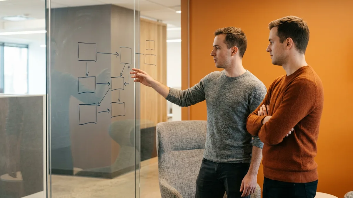 Two full stack developers sketching an architecture diagram of boxes and arrows on a glass whiteboard in an office with an orange accent wall