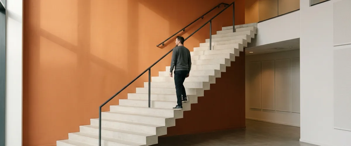 Engineer in a charcoal sweater climbing a wide concrete staircase beside a burnt-orange atrium wall in afternoon light