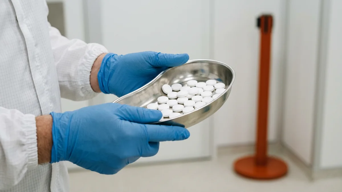 Blue-gloved hands holding a stainless steel tray of white tablets in a pharmaceutical cleanroom