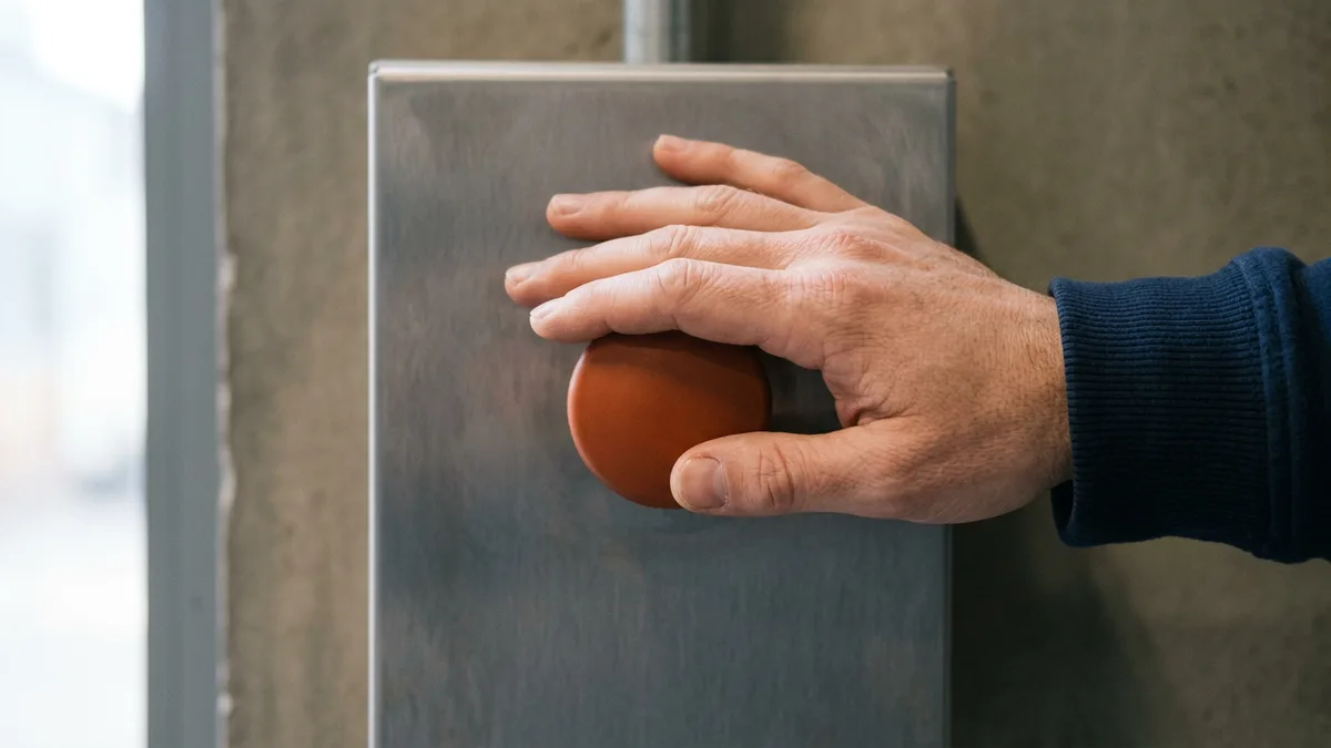 Hand resting on a plain orange stop button on a steel panel, representing who can switch off an AI feature