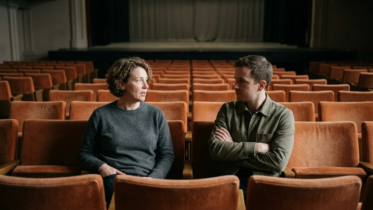 Two colleagues seated in orange velvet seats of an empty theater in conversation, illustrating a peer helping a cross-industry hire learn entertainment marketing