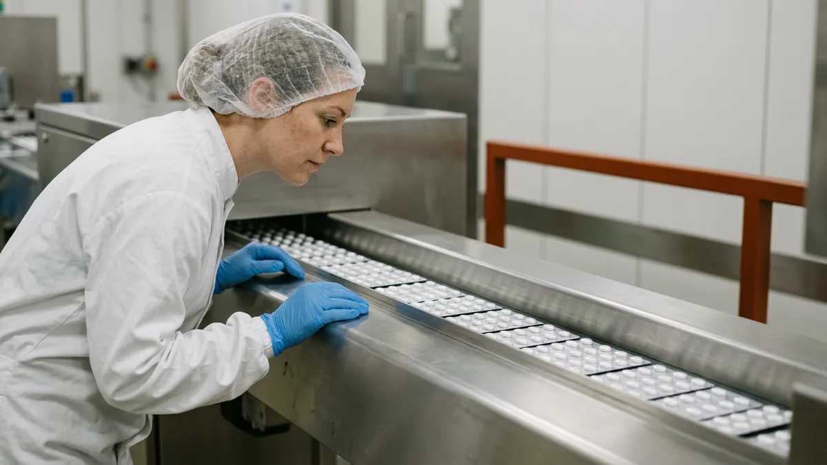 Operator in a hairnet and blue gloves inspecting blister packs of tablets on a pharmaceutical packaging line