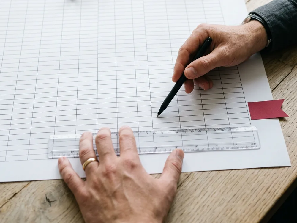 Two people tracing one row of a printed spreadsheet with a ruler and pen, with a red flag tab beside the row