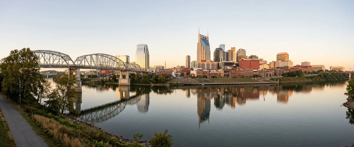 Downtown Nashville skyline across the Cumberland River at sunrise with a steel truss pedestrian bridge in the foreground