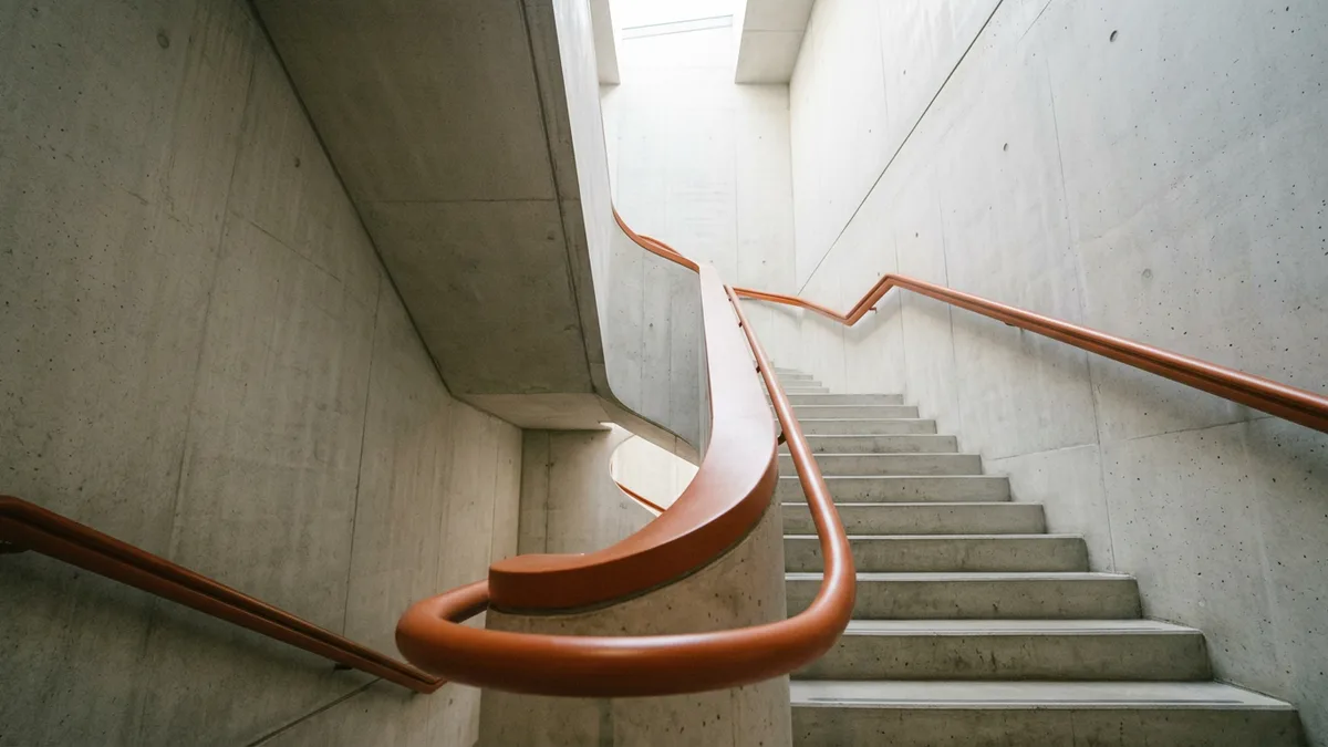 Orange handrail curving up a bare concrete office stairwell toward a skylight