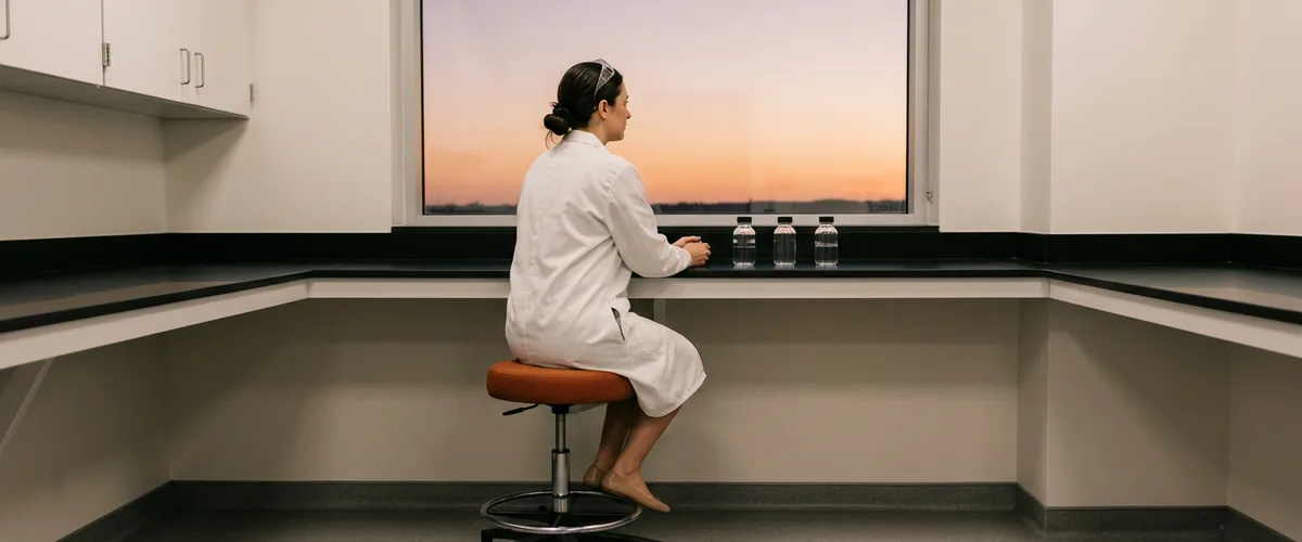 Quality control analyst in a white lab coat sitting at a black epoxy lab bench, looking out a window at dusk