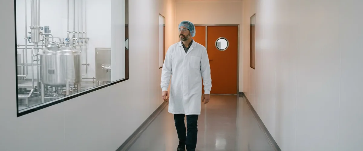 Quality manager in a white lab coat and hairnet walking a clean pharmaceutical plant corridor