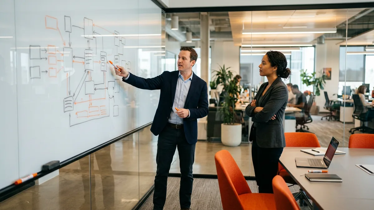 Two colleagues discussing a Splunk deployment architecture diagram sketched on a whiteboard in a glass conference room
