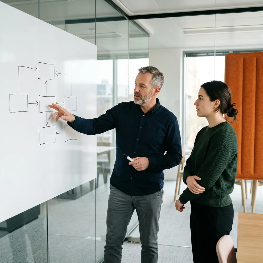 Two engineers at a whiteboard sketch of connected boxes and arrows, one explaining a design tradeoff while the other listens