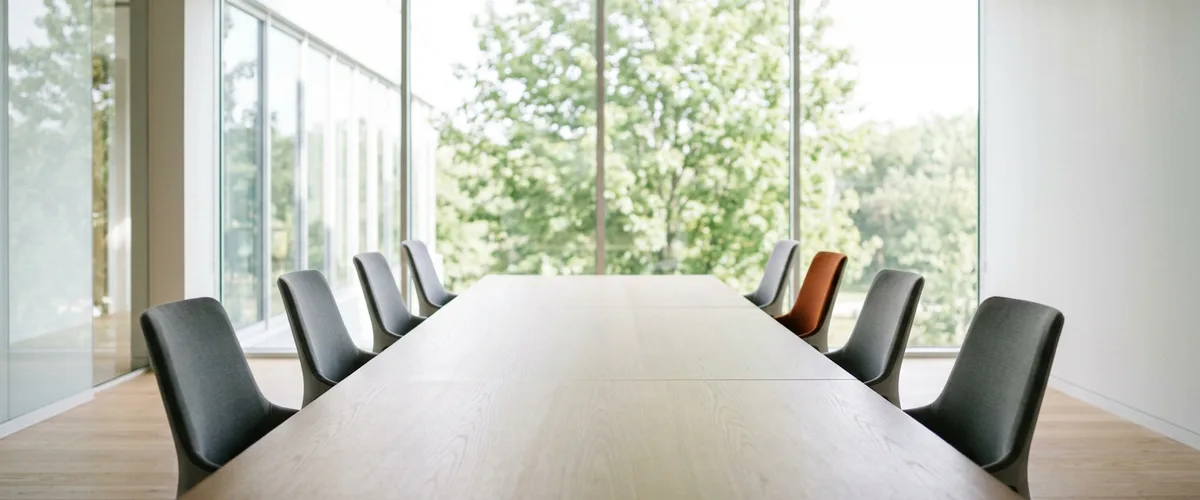 Long light-oak conference table with eight gray chairs and one burnt orange chair in a bright office with a window wall of trees