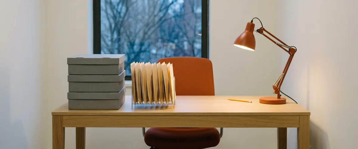 Empty accountant's desk at dawn with three gray archive boxes, a rack of manila folders, a pencil, and an orange desk lamp switched on