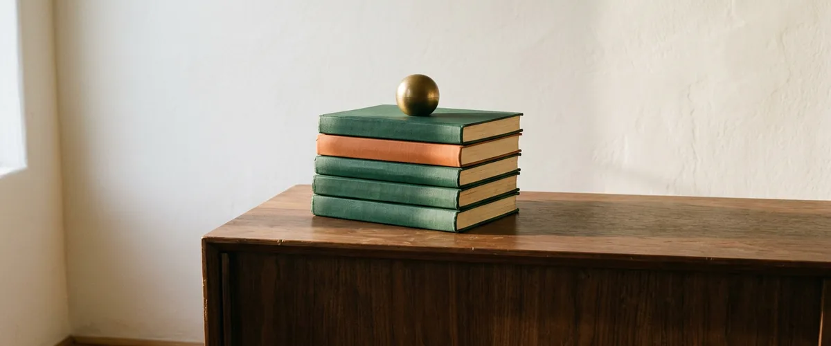 Stack of green and orange cloth-bound ledger books with a brass sphere paperweight on a walnut credenza in afternoon light