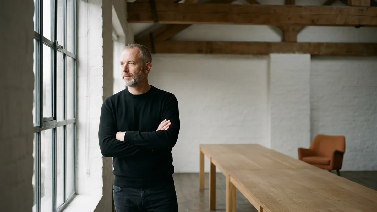 Creative director with a grey beard and black sweater standing with arms folded by a tall window in an agency loft with a long oak table