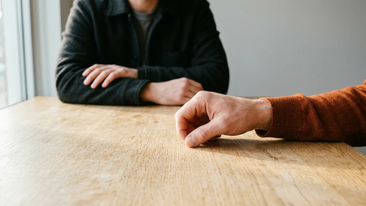 Hand in an orange sweater sleeve tapping its knuckles on an oak table while a listener in a dark jacket sits across with folded hands, illustrating the tappers and listeners experiment