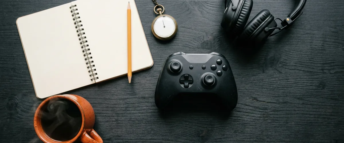 Black game controller beside an open blank notebook, a pencil, a pocket stopwatch, headphones and an orange coffee mug on a dark wooden desk
