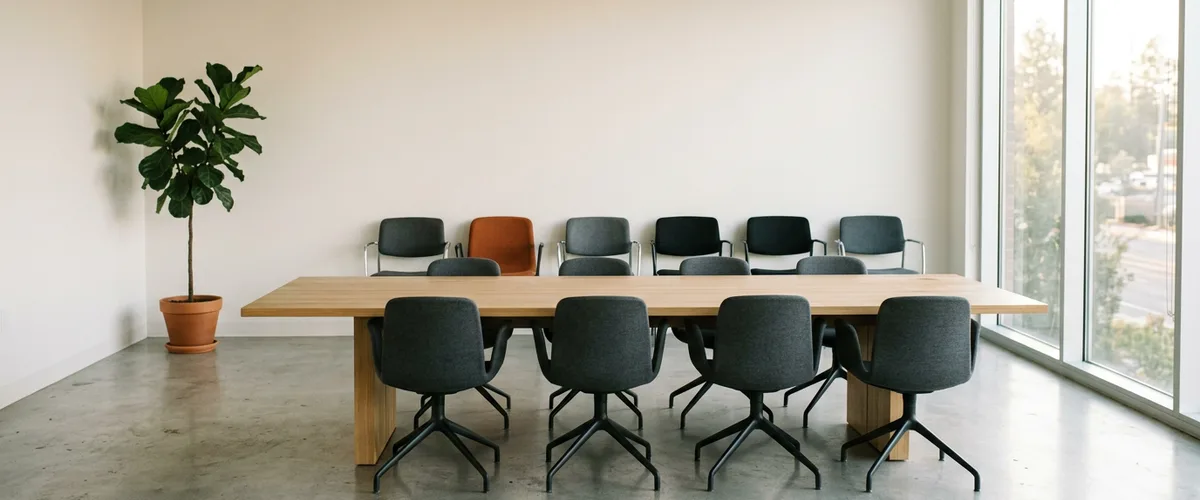 Empty conference room with eight gray chairs at a long oak table and a separate row of six chairs pushed back against the wall, one with a burnt orange seat