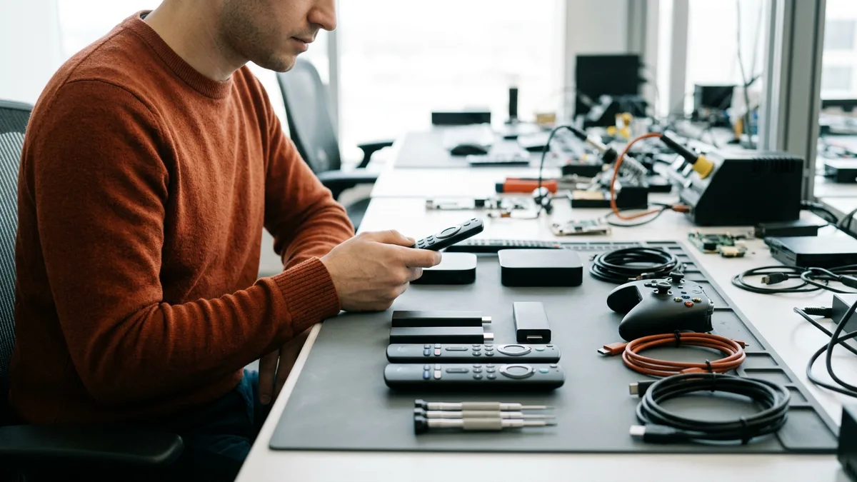 Streaming playback engineer in an orange sweater holding a remote control at a test bench of streaming sticks, set-top boxes, a game controller, and cables