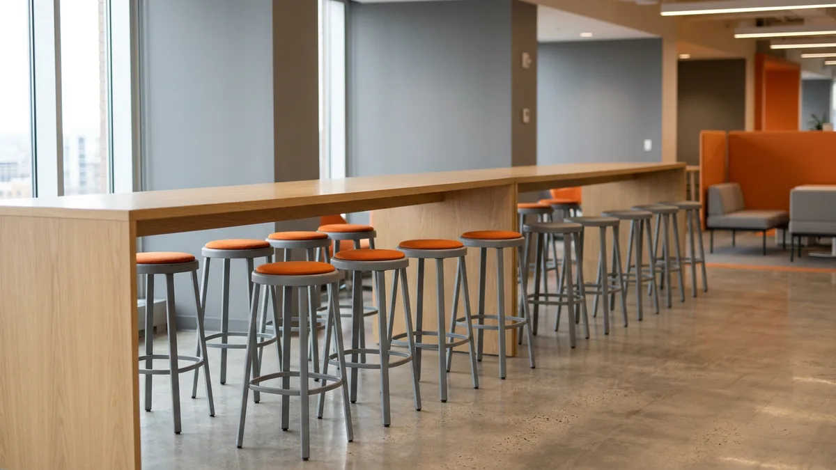 Row of gray bar stools along a long oak counter in a bright office, the nearer stools topped with orange cushions and the far ones bare
