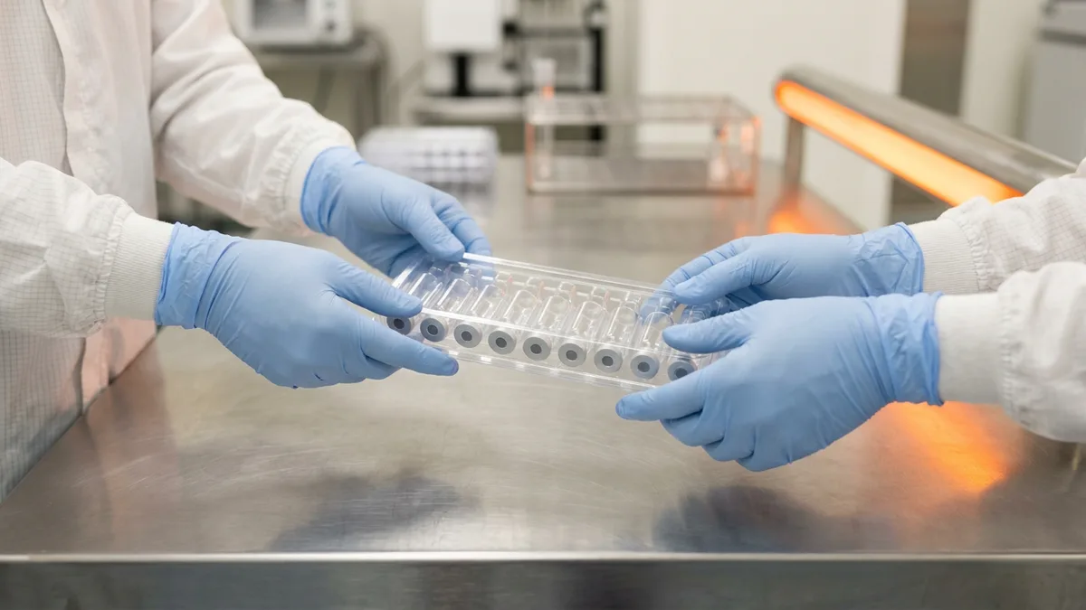 Two pairs of gloved hands passing a tray of glass vials across a stainless steel bench