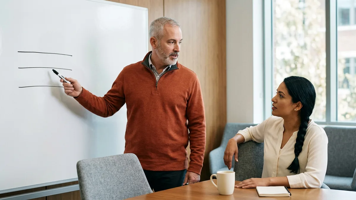 Interviewer in an orange quarter-zip pointing at three horizontal lines on a whiteboard while a colleague with a braid listens at the table
