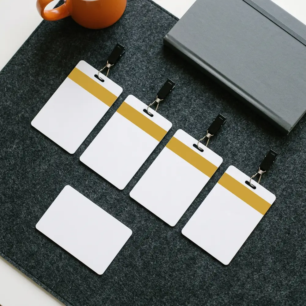 Four blank white ID badges with yellow contractor bands and one plain white badge laid out on a charcoal desk mat beside a grey notebook and an orange mug